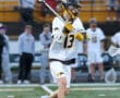 Lacrosse player wearing white jersey throws ball during game, with spectators in the background.