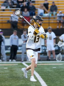 Lacrosse player wearing white jersey throws ball during game, with spectators in the background.