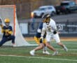 High school lacrosse game action with a player dodging an opponent near the goal. Team jerseys in vibrant colors.