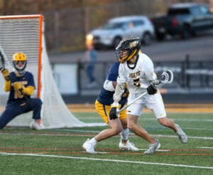 High school lacrosse game action with a player dodging an opponent near the goal. Team jerseys in vibrant colors.