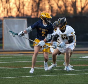 Lacrosse players from Mt. Lebanon and Tigers compete for the ball on a field during a dramatic sunset.