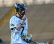 Lacrosse player wearing a helmet and jersey number 7, smiling while holding a stick on the field.