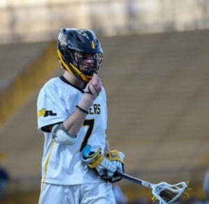 Lacrosse player wearing a helmet and jersey number 7, smiling while holding a stick on the field.