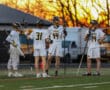 Four lacrosse players in team uniforms discuss strategy during practice, with a vibrant sunset backdrop.