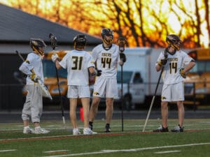 Four lacrosse players in team uniforms discuss strategy during practice, with a vibrant sunset backdrop.