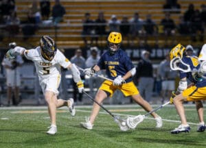 Two high school lacrosse players compete for the ball during a night game on a well-lit field.