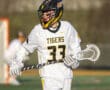 Lacrosse player wearing a Tigers jersey, helmet, and gloves, running on the field with a lacrosse stick.