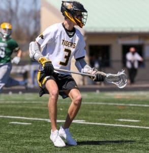 Lacrosse player in a white jersey with Tigers on it, running on the field while holding a lacrosse stick.