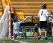 Goalie makes a save during a lacrosse game, with teammates in the background and empty stands visible.