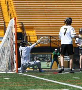 Goalie makes a save during a lacrosse game, with teammates in the background and empty stands visible.