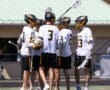 Lacrosse players in team jerseys huddle together, strategizing during a game on a sunny field.