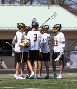 Lacrosse players in team jerseys huddle together, strategizing during a game on a sunny field.