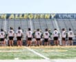 Lacrosse players from North Allegheny and St. Edward prepare for the game at the field with empty bleachers in the background.