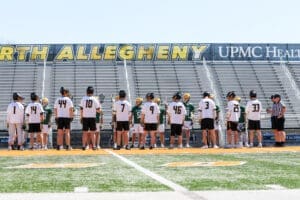 Lacrosse players from North Allegheny and St. Edward prepare for the game at the field with empty bleachers in the background.