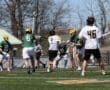 Lacrosse game in action, featuring players in green and white jerseys on a sunny day, with a referee officiating.