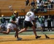 Lacrosse players compete on the field, showcasing athleticism during a game, with fans watching in the background.