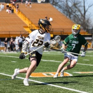 Two young lacrosse players compete on a sunny field, one in a white Tigers jersey and the other in a green St. Edward jersey.