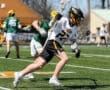 High school lacrosse match with a player in a black uniform sprinting while being closely guarded by an opponent.