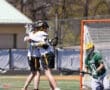 Lacrosse players celebrate a goal with a hug while a goalie watches from the net during a sunny game day.