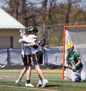Lacrosse players celebrate a goal with a hug while a goalie watches from the net during a sunny game day.