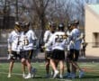 Lacrosse team celebrating on the field, wearing white and black uniforms, in a sunny outdoor setting.