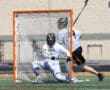 Lacrosse game action, goalie in white and black uniform defending against a shot with an orange goal in focus.