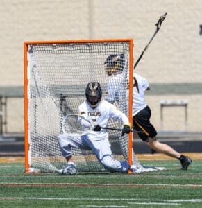 Lacrosse game action, goalie in white and black uniform defending against a shot with an orange goal in focus.