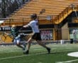 High school lacrosse game action, with players in motion on a green field and stadium seating in the background.