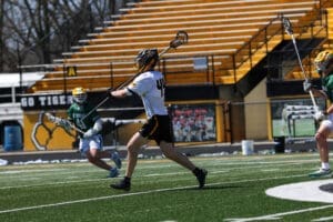 High school lacrosse game action, with players in motion on a green field and stadium seating in the background.