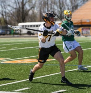 Lacrosse players compete on a sunny day, showcasing skills on the field during a match. Player in focus wears number 7.
