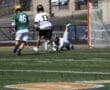 Lacrosse players competing on the field, one diving for the ball while others pursue, with a goal in the background.