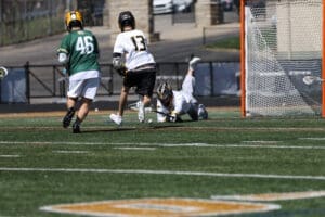Lacrosse players competing on the field, one diving for the ball while others pursue, with a goal in the background.