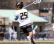 Lacrosse player in action, wearing jersey number 25, practicing a shot on a sunny field with teammates in the background.