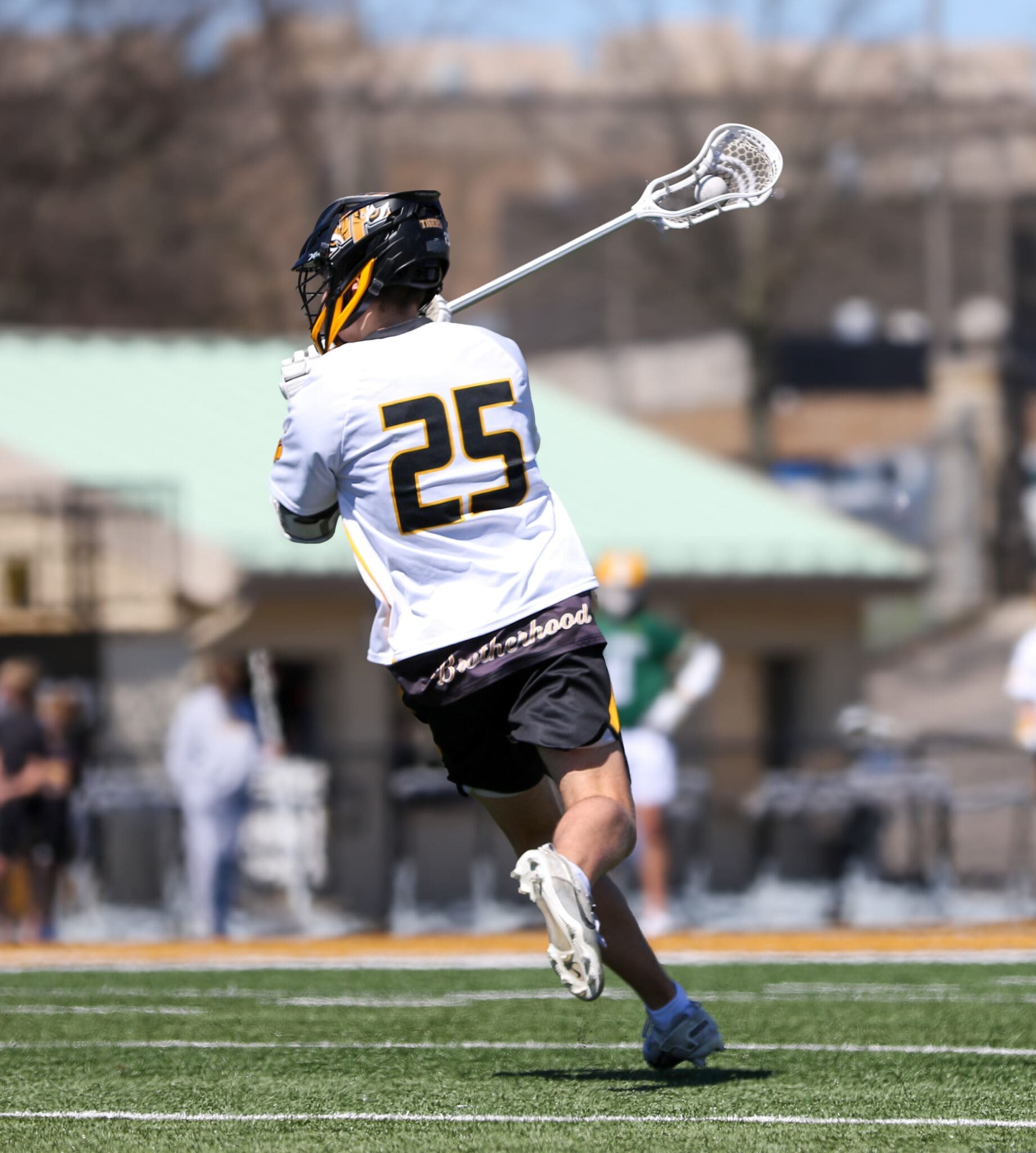 Lacrosse player in action, wearing jersey number 25, practicing a shot on a sunny field with teammates in the background.