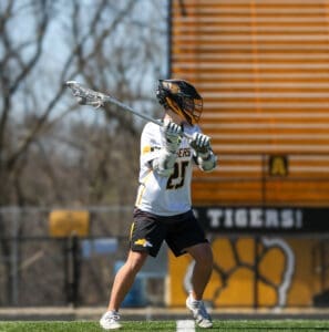 Lacrosse player in action on the field, ready to pass, wearing a helmet and team jersey with Tigers logo.