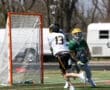 Lacrosse players compete on the field; one shoots while the other defends the goal. Energy and action captured in motion.