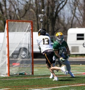 Lacrosse players compete on the field; one shoots while the other defends the goal. Energy and action captured in motion.