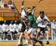 Action shot of lacrosse players competing fiercely on the field during a sunny day, with spectators in the background.