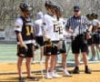 Lacrosse players in jerseys await instructions from a referee during a game on a sunny day. Team uniforms feature tiger logos.
