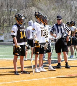 Lacrosse players in jerseys await instructions from a referee during a game on a sunny day. Team uniforms feature tiger logos.