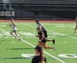 Lacrosse players sprinting across a green field during a game, with a referee overseeing the action.