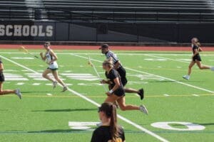Lacrosse players sprinting across a green field during a game, with a referee overseeing the action.