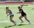 Two female lacrosse players compete on a bright green turf field during an intense game, wearing protective gear.