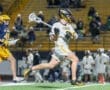 High school lacrosse players in action, one running with a stick while the other pursues on the field at night.