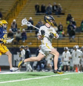 High school lacrosse players in action, one running with a stick while the other pursues on the field at night.