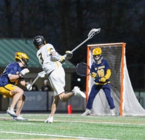 Lacrosse player in motion takes a shot on goal, with a goalie poised to defend during an evening match on a turf field.