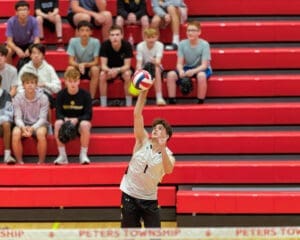 High school volleyball player preparing to serve a volleyball in front of an audience on red bleachers.