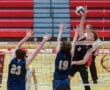 High school volleyball match action with a player spiking the ball over defenders at the net in an indoor gymnasium.