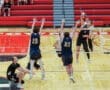 High school volleyball players compete at the net, with one player spiking the ball during an intense match.