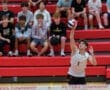 High school volleyball player serves the ball, with enthusiastic teammates cheering in the background on red bleachers.